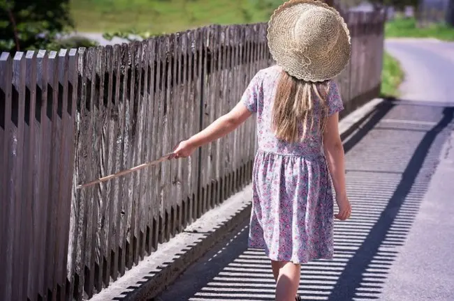Девочка в шляпке идет по дороге и ведет палкой по забору для асмр звуков