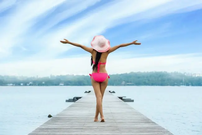 Девушка в розовой шляпке и красном купальнике на берегу озера раскинула руки в радости